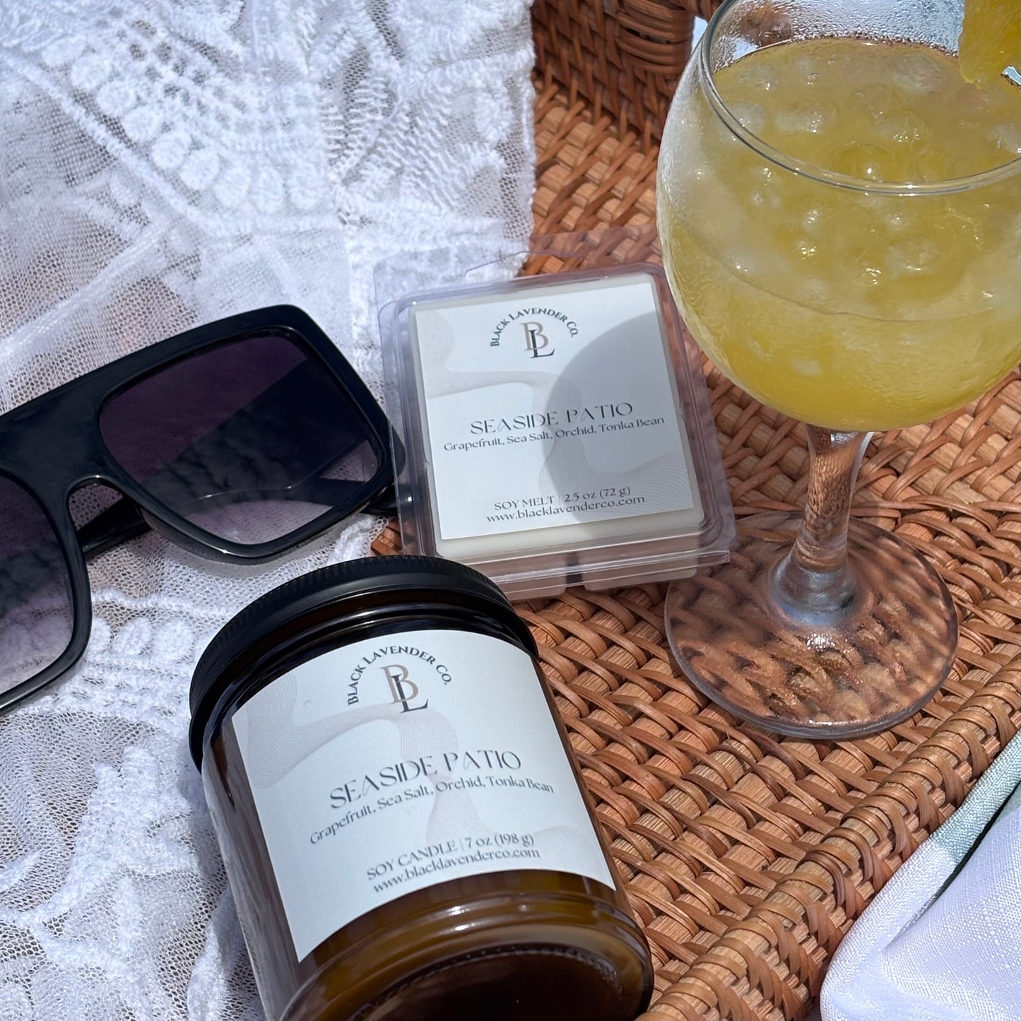 Candle, wax melts, sunglasses, and a glass of yellow drink on a woven surface.