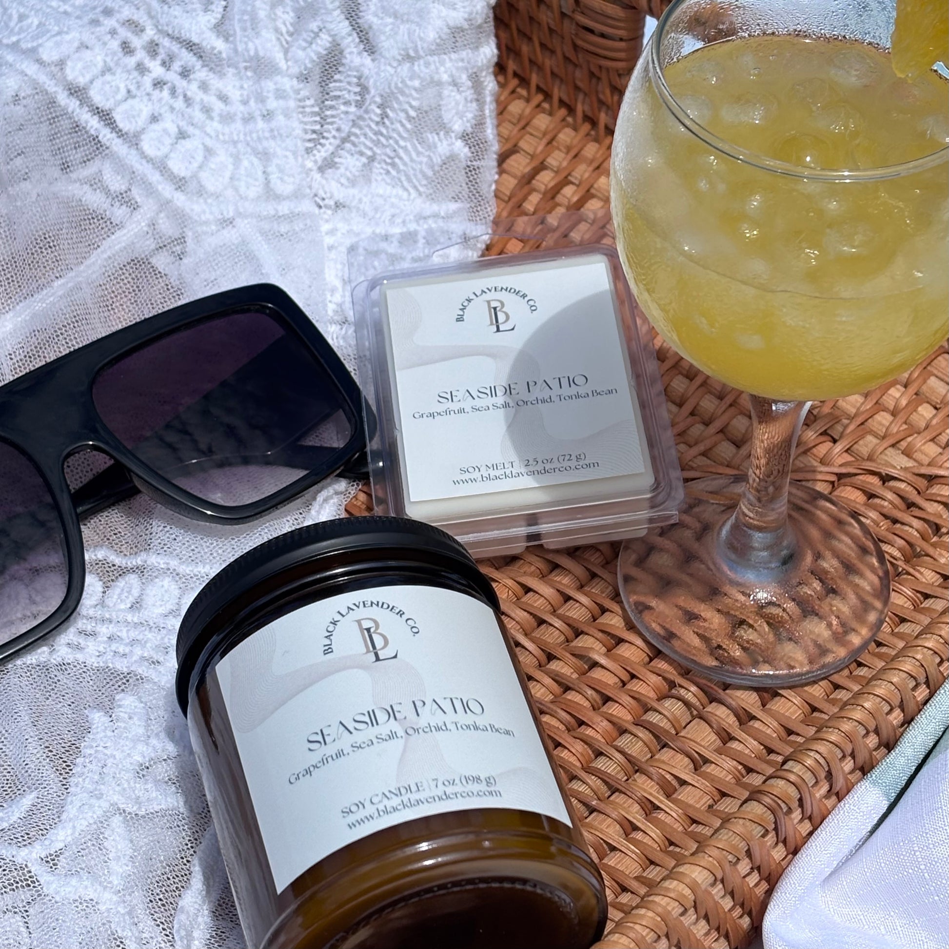 Candle, wax melts, sunglasses, and a glass of yellow drink on a woven surface.
