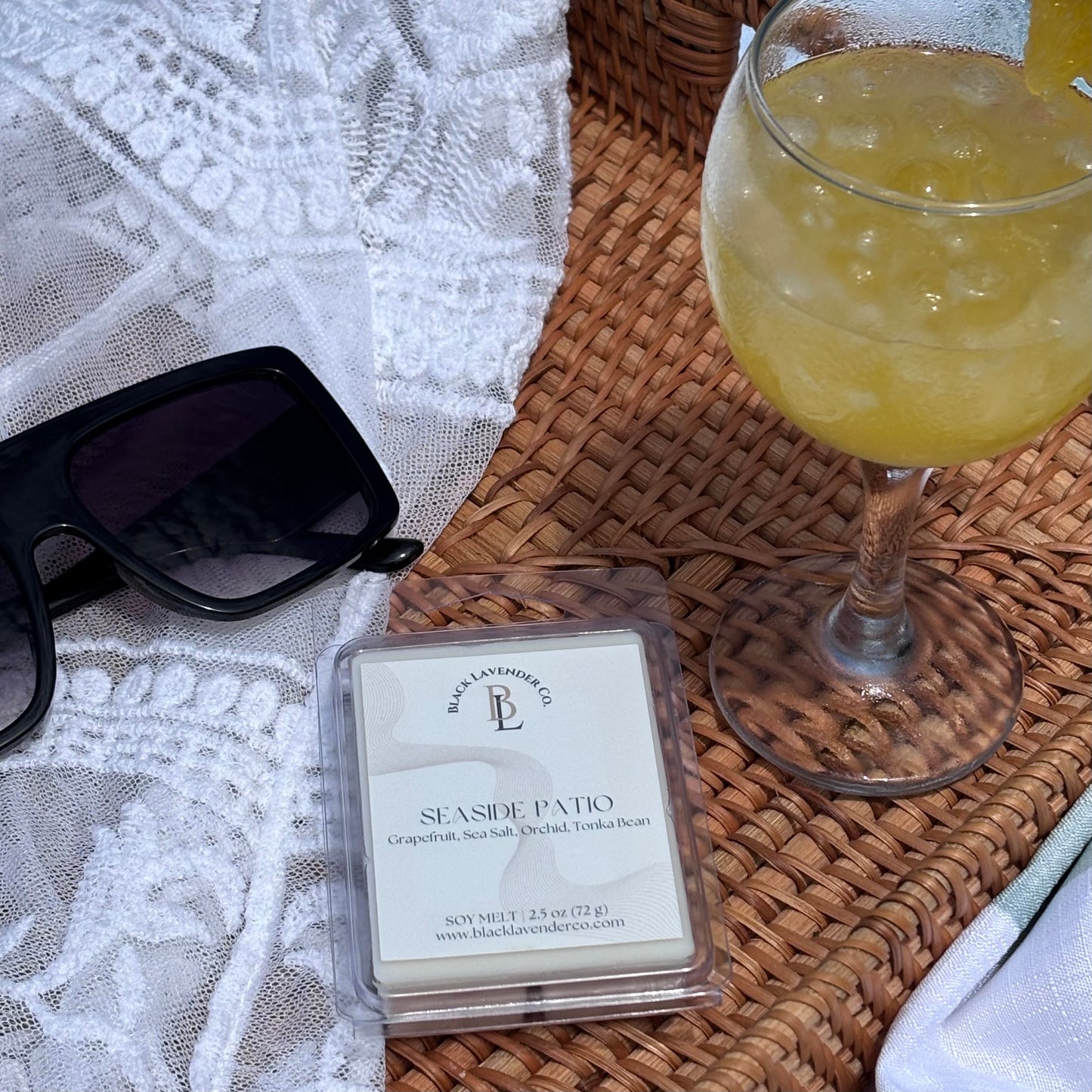 Wax melt packaging labeled 'Seaside Patio' next to a glass of yellow drink on a woven surface.