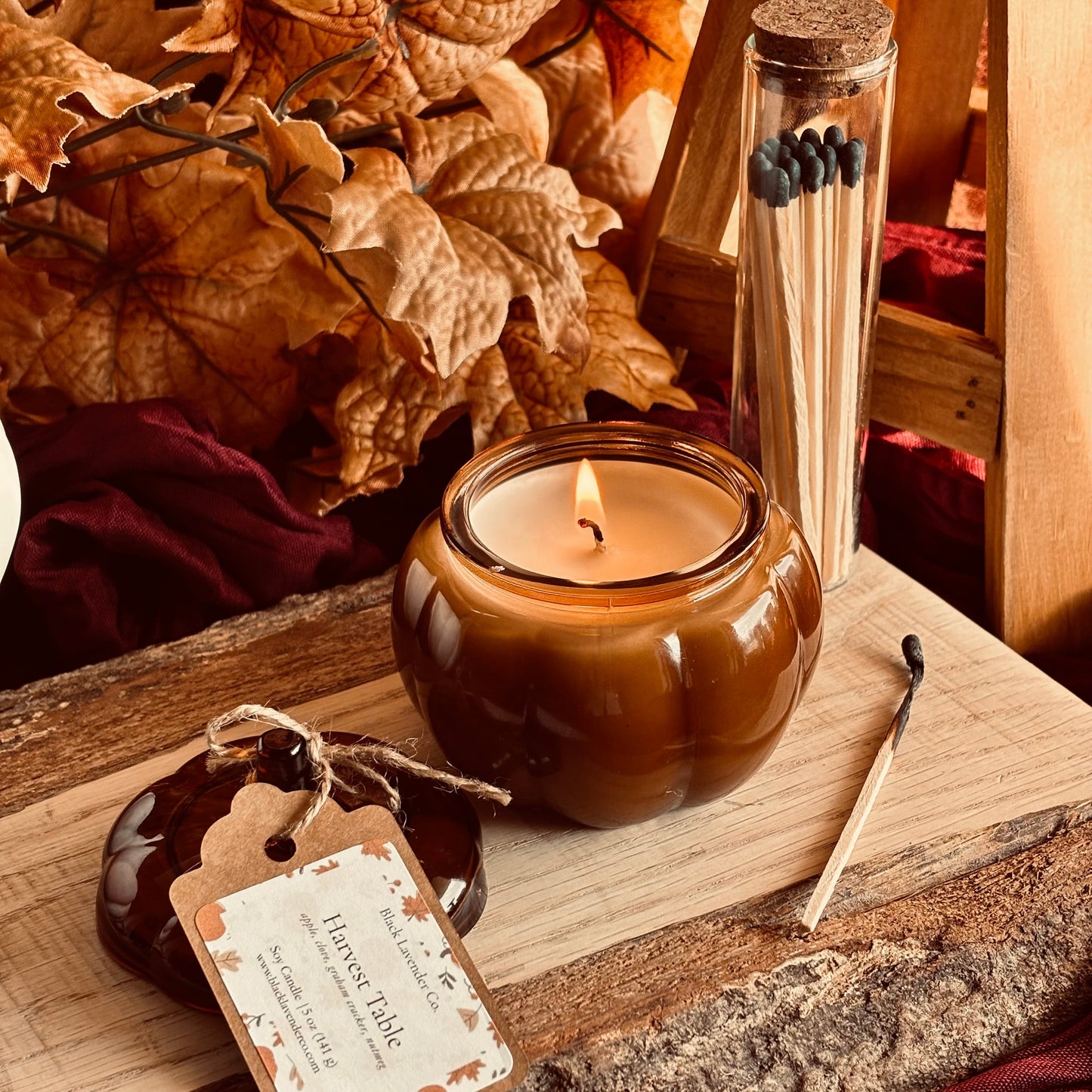 A lit amber glass candle jar sits on a rustic wood surface with a matching tag, a single match, and a glass tube of matches in a warm autumn-themed setting with dried leaves and burgundy fabric.
