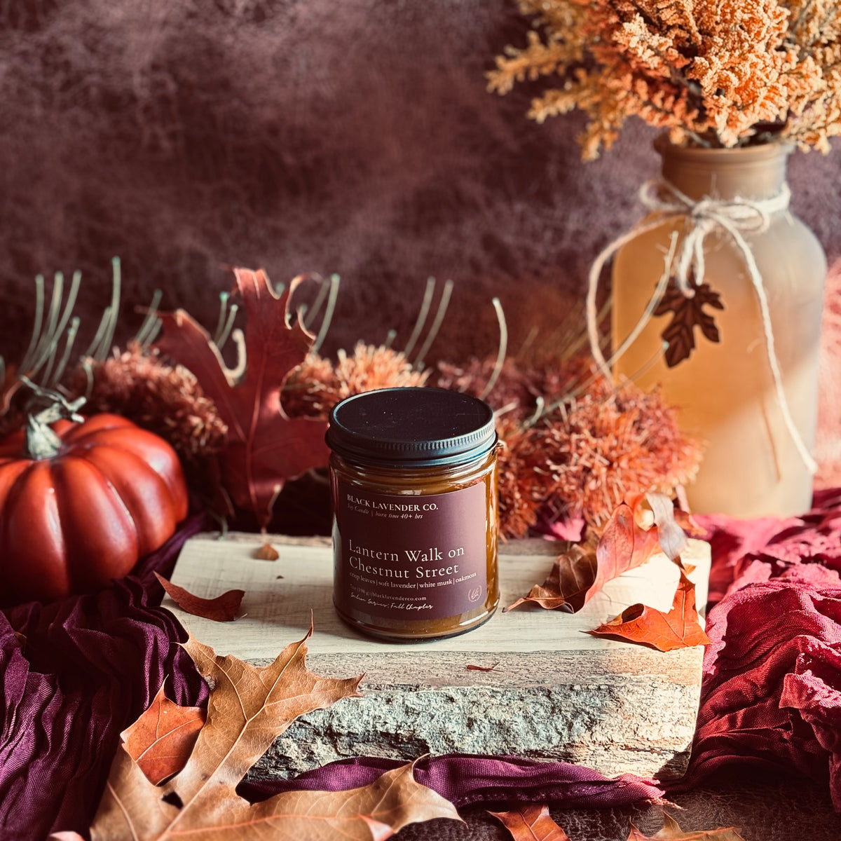 A candle jar from Black Lavender Co. styled in an autumn scene with a small pumpkin, dried flowers, and leaves on a rustic wooden surface.