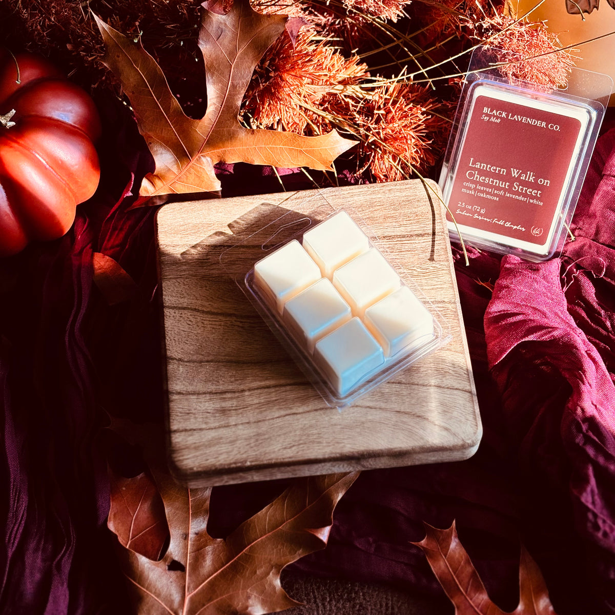 A wax melt product by Black Lavender Co. displayed on a wooden block with autumn-themed props including a small pumpkin, dried leaves, and burgundy fabric.