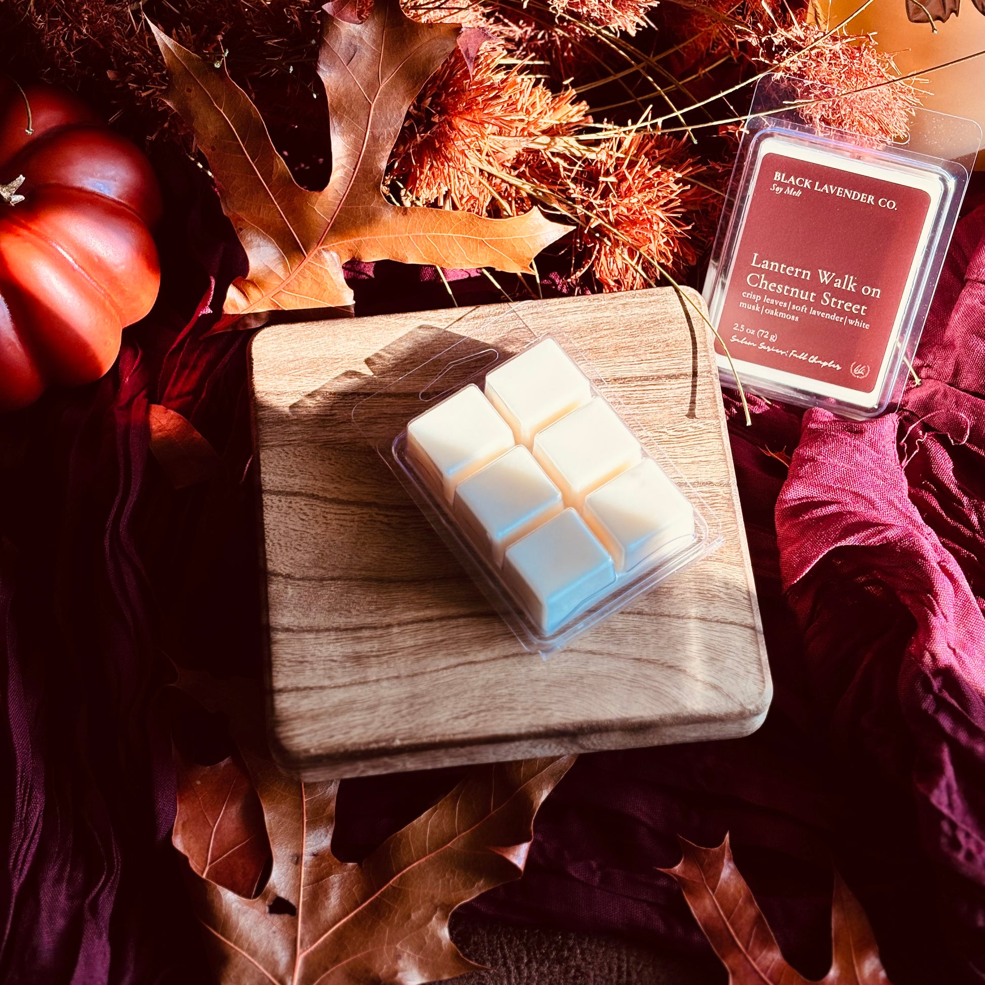 A wax melt product by Black Lavender Co. displayed on a wooden block with autumn-themed props including a small pumpkin, dried leaves, and burgundy fabric.
