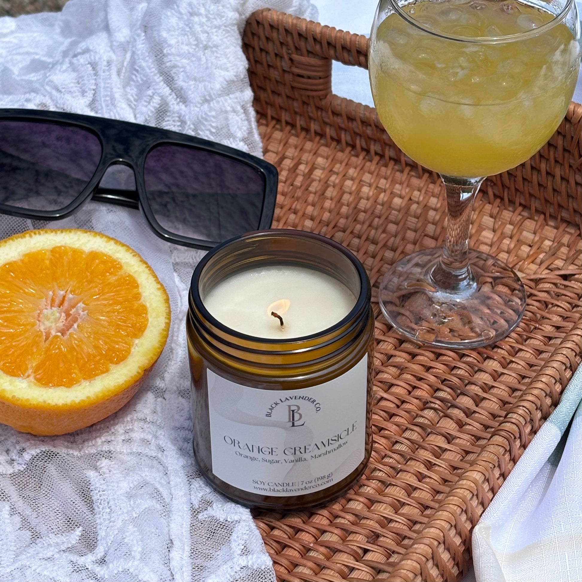 Candle labeled 'Orange Creamsicle' next to a glass of orange juice and half an orange on a woven tray.