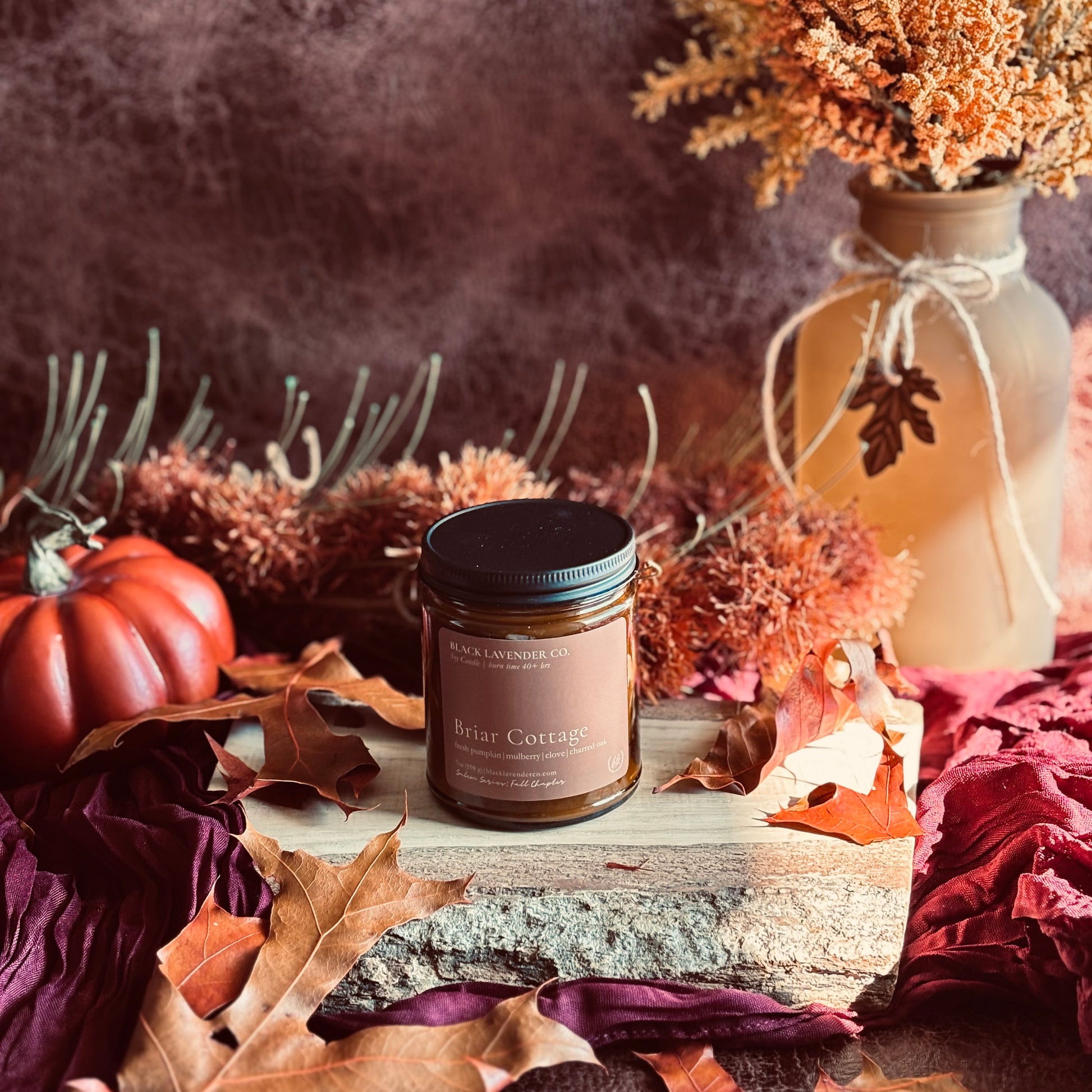 A jar candle labeled 'Briar Cottage' by 'Black Lavender Co.' is styled on a rustic wood slab with autumn leaves, a small orange pumpkin, dried flowers in a vase, and a smoky brown background.