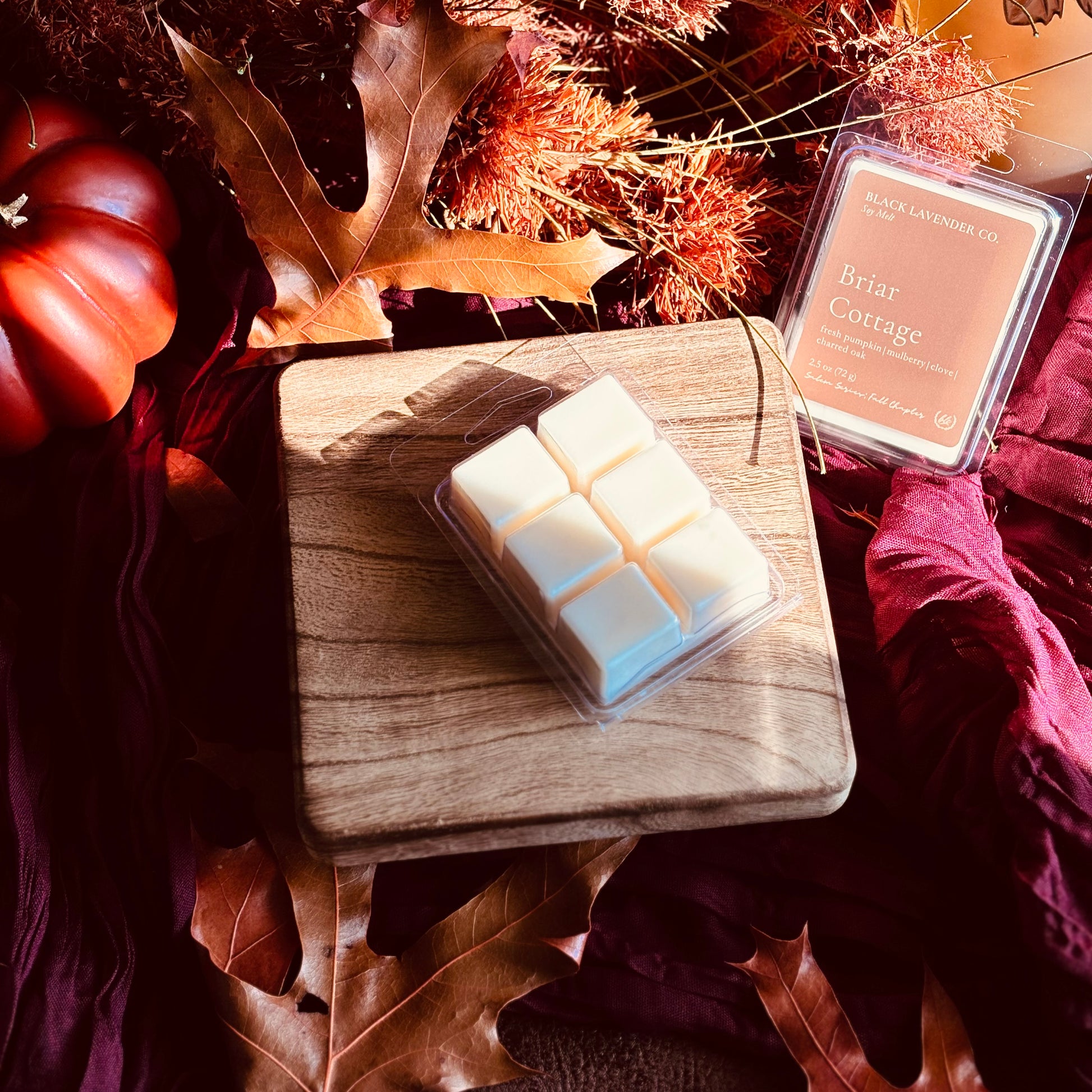 A wax melt product set from Black Lavender Co. featuring a six-cell white wax melt on a wooden block and its clear plastic clamshell container labeled 'Briar Cottage' in a cozy autumn setting with pumpkins, leaves, and draped fabric.