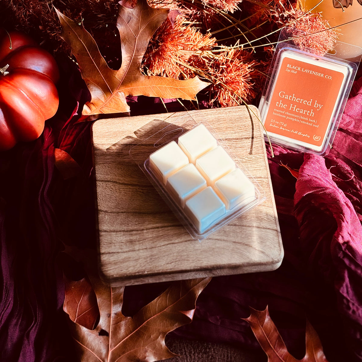 A wax melt product displayed on a small wooden board, with its branded clamshell package beside it and autumn leaves, fabric, and a small pumpkin used as styled props.
