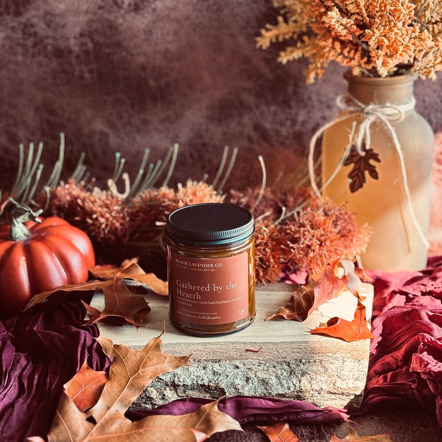 A jar candle labeled 'Gathered by the Hearth' by Black Lavender Co sits on a rustic wood slab surrounded by autumn leaves, a small orange pumpkin, dried flowers in a vase, and a dark warm-toned background.