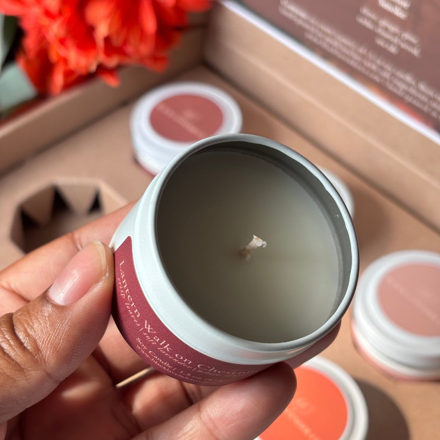 A hand holds a small open candle tin with pale wax and a pink label, with other similar candles and a decorative orange flower in the background.