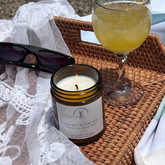 Candle labeled 'Coastal Escape' with a glass of yellow liquid and sunglasses on a wicker tray.