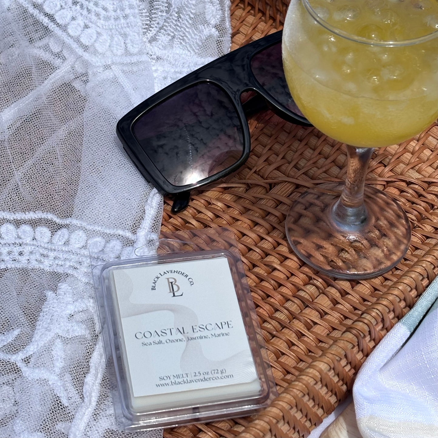 Black Lava Candle Co. 'Coastal Escape' wax melt with sunglasses and a cocktail on a woven surface.