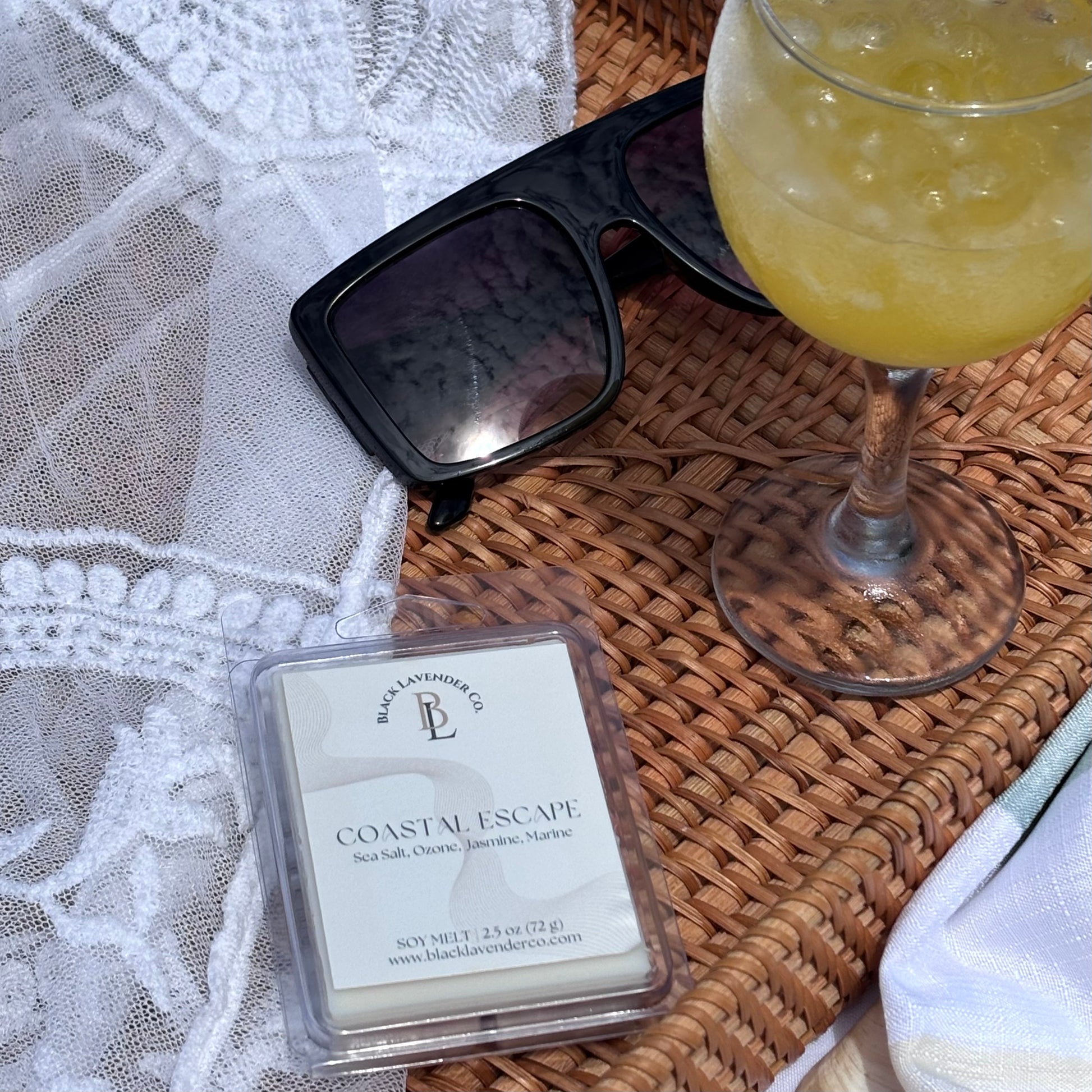 Black Lava Candle Co. 'Coastal Escape' wax melt with sunglasses and a cocktail on a woven surface.