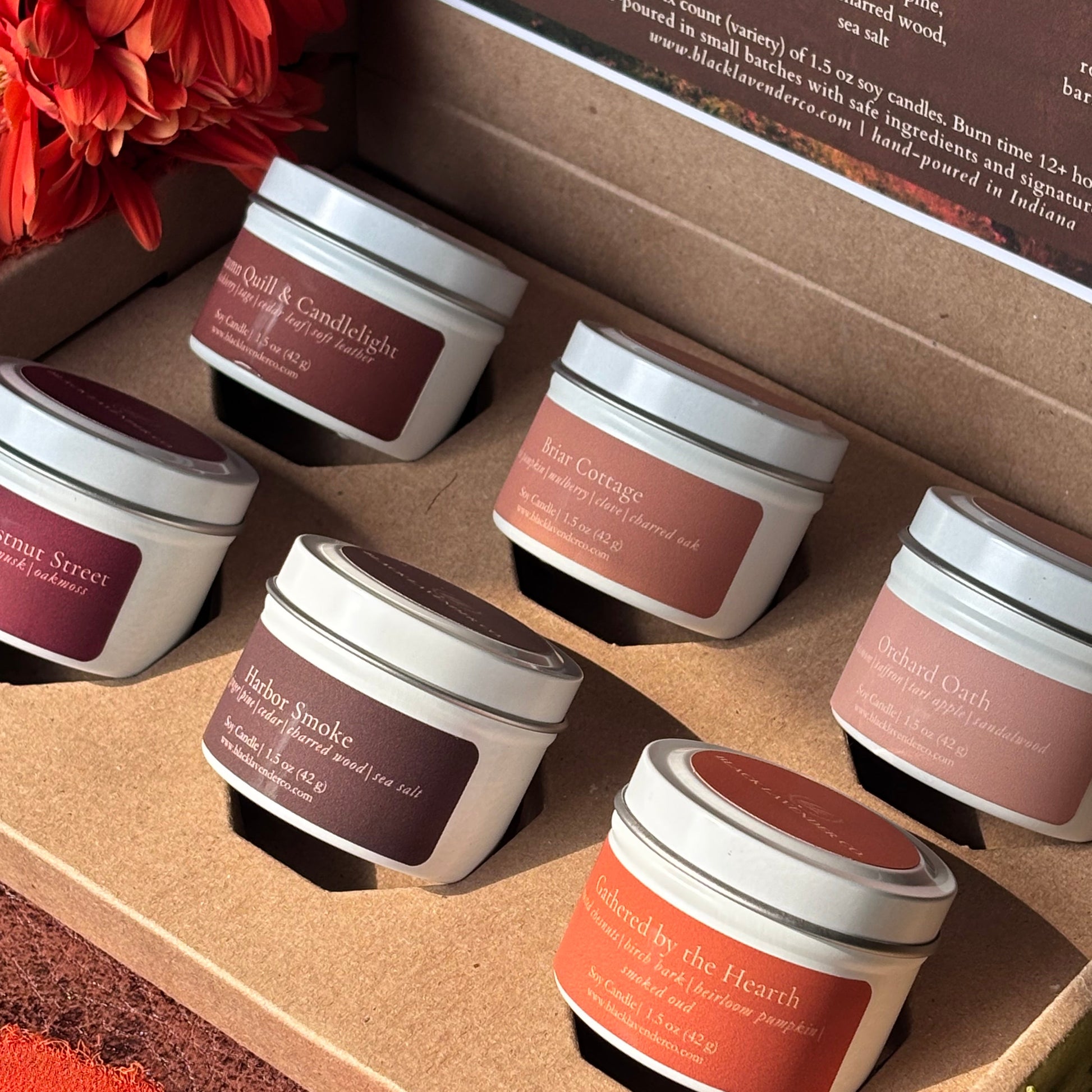Six small white jars of scented candles arranged in a cardboard tray, each with a different warm-toned label in shades of brown, terracotta, and dusty rose. A large red flower arrangement sits in the upper left corner, and part of a printed card is visible inside the box.
