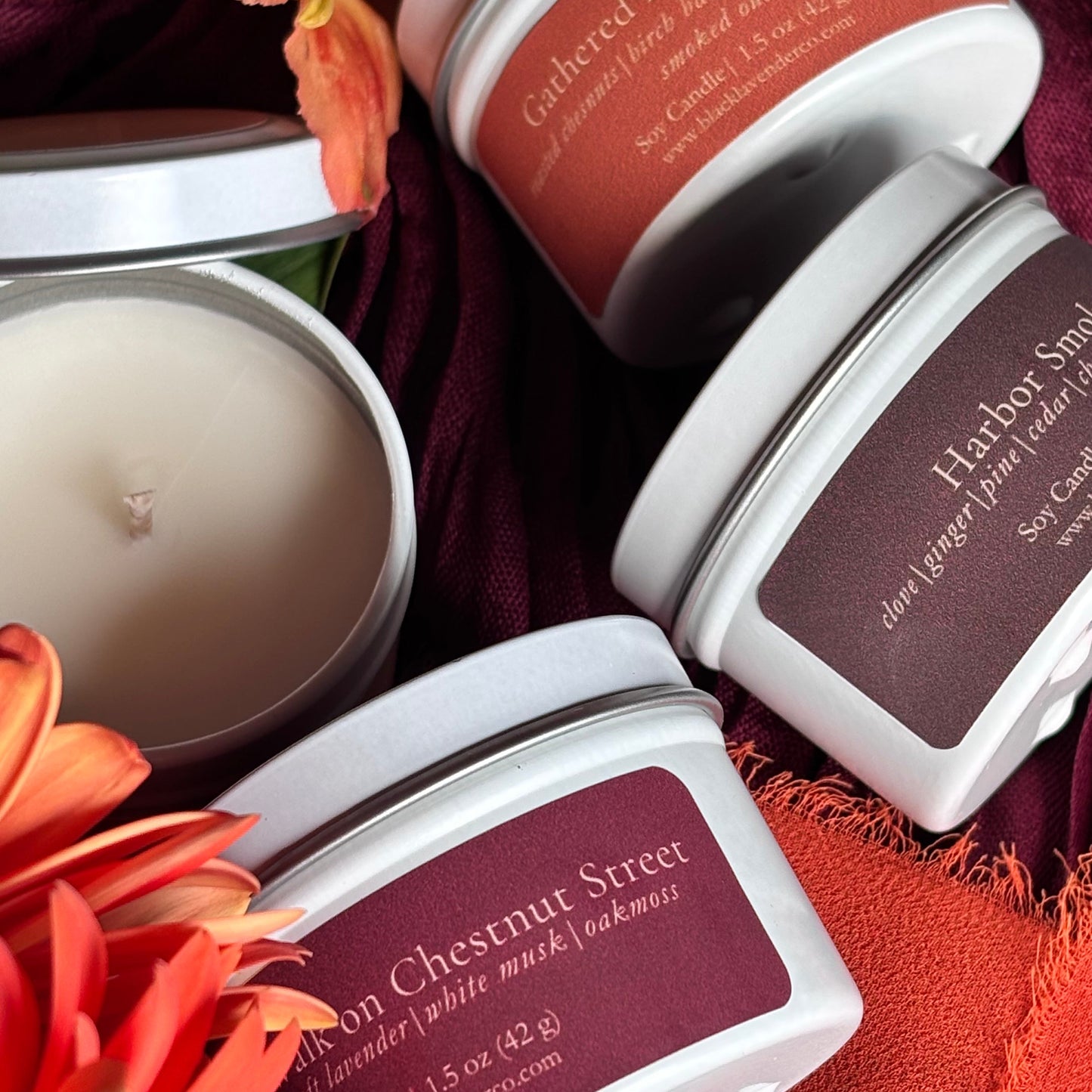 Three white metal tins of scented candles arranged on a dark burgundy fabric with orange flowers. One tin is open, showing the cream-colored wax.