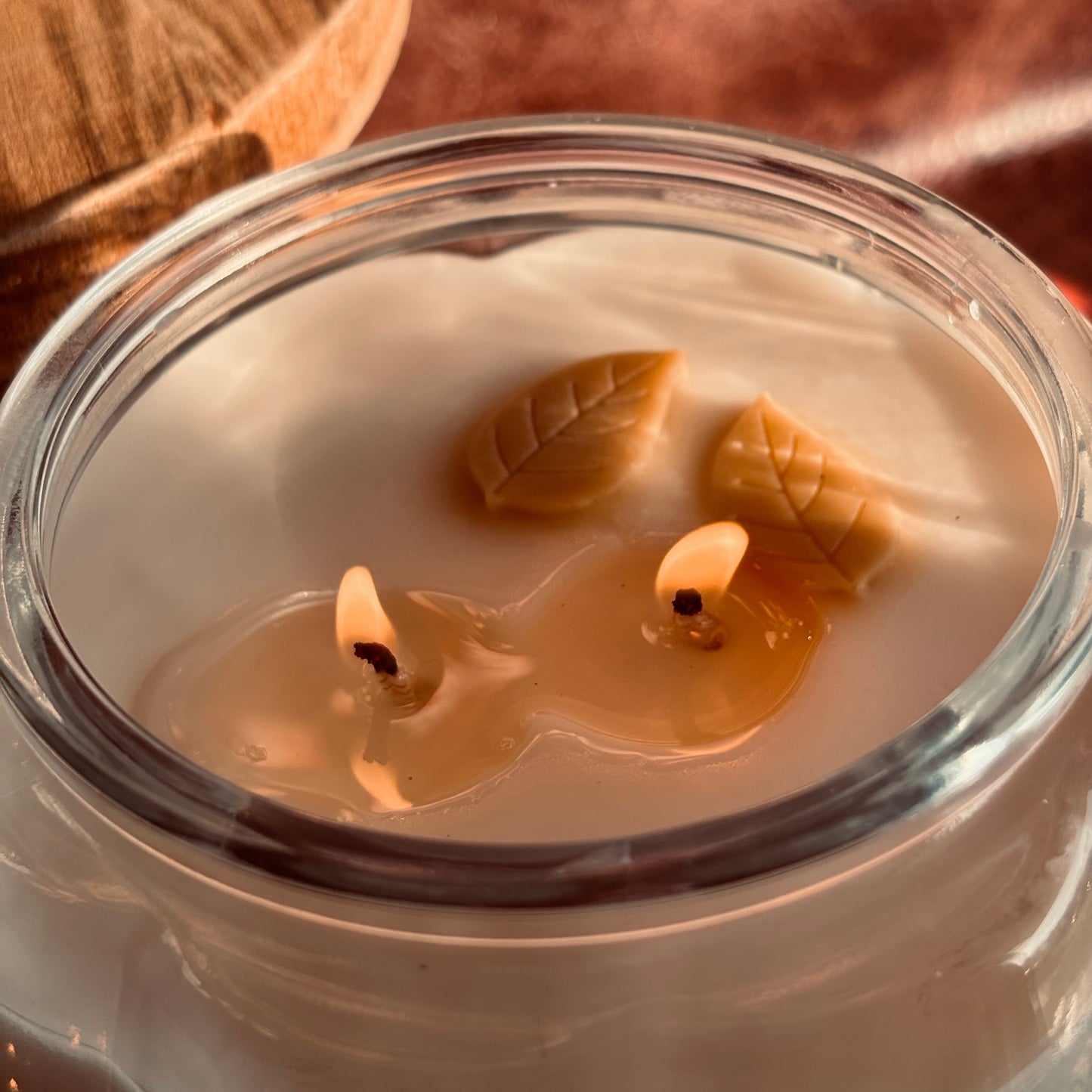 A close-up of a lit soy wax candle in a clear glass jar, featuring two wicks and decorative leaf-shaped wax ornaments floating on the surface. The warm glow of the flames illuminates the cream-colored wax, with a softly blurred wooden background.